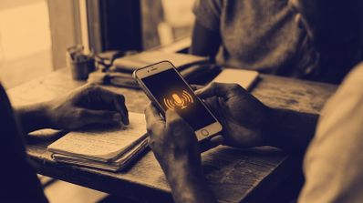 Manos sosteniendo un smartphone que muestra un icono de micrófono y ondas de sonido durante una conversación en una mesa.