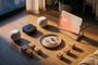 Conjunto de dispositivos Apple en el suelo, incluyendo HomePod mini, Apple TV y una pantalla inteligente experimental, rodeados de iconos de domótica grabados en madera.