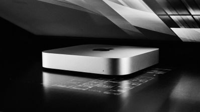 Mac mini M6 de color plata sobre una superficie oscura bajo la luz de un monitor.