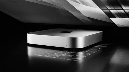 Mac mini M6 de color plata sobre una superficie oscura bajo la luz de un monitor.