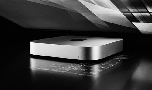 Mac mini M6 de color plata sobre una superficie oscura bajo la luz de un monitor.