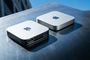 Dos ordenadores Mac mini colocados sobre una superficie metálica, uno mostrando su panel frontal y el otro su parte trasera con puertos de conexión.