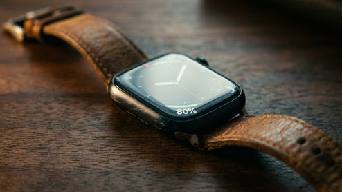 Primer plano de un Apple Watch Series 8 con correa de cuero marrón sobre una mesa de madera, mostrando un 80% de batería en pantalla.