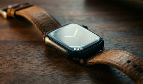 Primer plano de un Apple Watch Series 8 con correa de cuero marrón sobre una mesa de madera, mostrando un 80% de batería en pantalla.