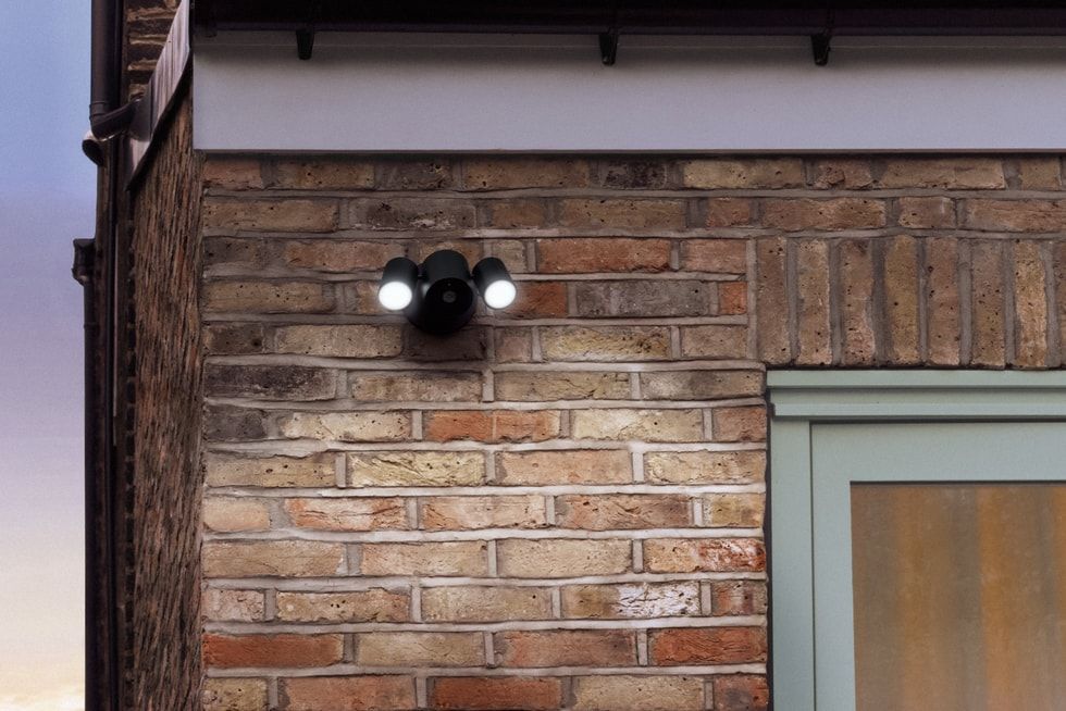 Cámara de seguridad inteligente de color negro con dos focos LED encendidos instalada en una fachada de ladrillo visto.