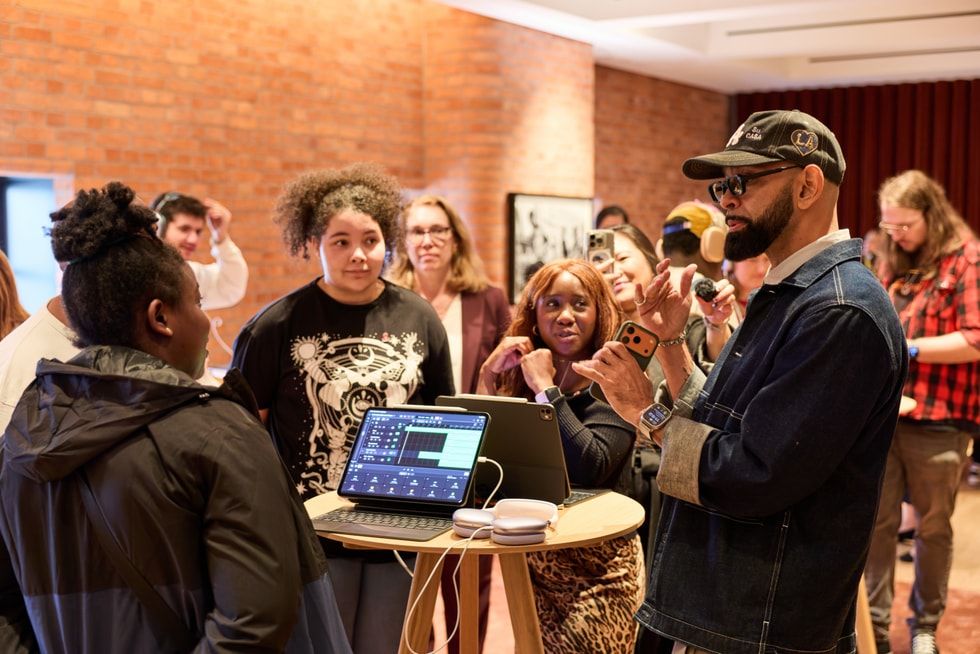 Un instructor con gorra y chaqueta vaquera gesticula frente a un grupo de jóvenes mientras explica el uso de una aplicación de edición musical en un iPad.