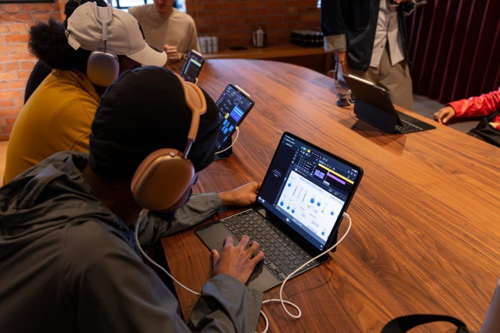 Varios jóvenes utilizan iPads con auriculares y teclados para trabajar en edición de audio y producción musical sobre una mesa de madera.