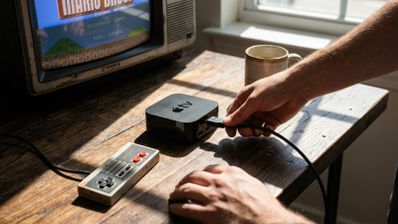 Persona conectando un cable a un Apple TV 2 junto a un mando de NES y una televisión antigua que muestra un videojuego clásico.