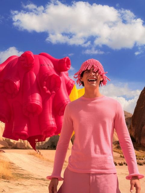Modelo masculino con ropa y gorro de cuentas rosa frente a una estructura inflable fucsia en un paisaje desértico.