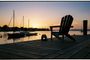Silla de madera y prismáticos en un muelle al atardecer con veleros fondeados al fondo.