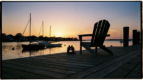 Silla de madera y prismáticos en un muelle al atardecer con veleros fondeados al fondo.