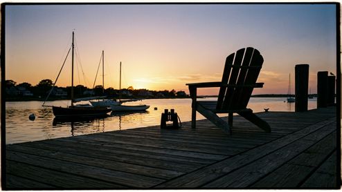 Silla de madera y prismáticos en un muelle al atardecer con veleros fondeados al fondo.