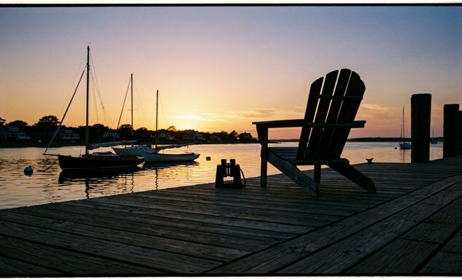 Silla de madera y prismáticos en un muelle al atardecer con veleros fondeados al fondo.