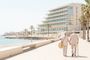 Pareja de jubilados caminando de la mano por un paseo marítimo frente a un hotel moderno y la playa.