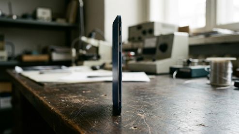 Vista de perfil de un smartphone extremadamente fino de color azul oscuro colocado verticalmente sobre una mesa de trabajo de madera en un taller.