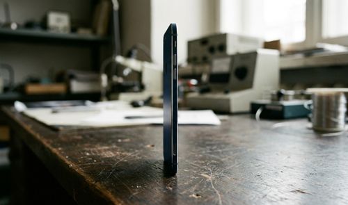 Vista de perfil de un smartphone extremadamente fino de color azul oscuro colocado verticalmente sobre una mesa de trabajo de madera en un taller.