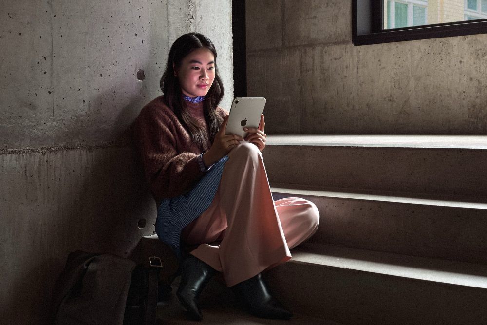 Una mujer joven sentada en unas escaleras de hormigón mientras utiliza un iPad mini de Apple en un entorno de luz tenue.