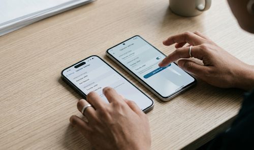 Una persona gestiona simultáneamente los ajustes de transferencia de eSIM en un iPhone y un smartphone Android situados sobre una mesa de madera.