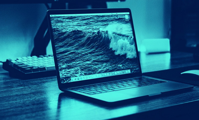 Ordenador portátil MacBook Pro abierto sobre un escritorio con un fondo de pantalla que muestra una ola rompiendo en el mar, bajo una iluminación en tonos azules.