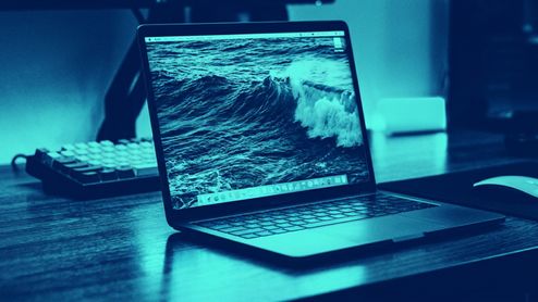 Ordenador portátil MacBook Pro abierto sobre un escritorio con un fondo de pantalla que muestra una ola rompiendo en el mar, bajo una iluminación en tonos azules.