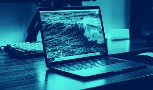 Ordenador portátil MacBook Pro abierto sobre un escritorio con un fondo de pantalla que muestra una ola rompiendo en el mar, bajo una iluminación en tonos azules.